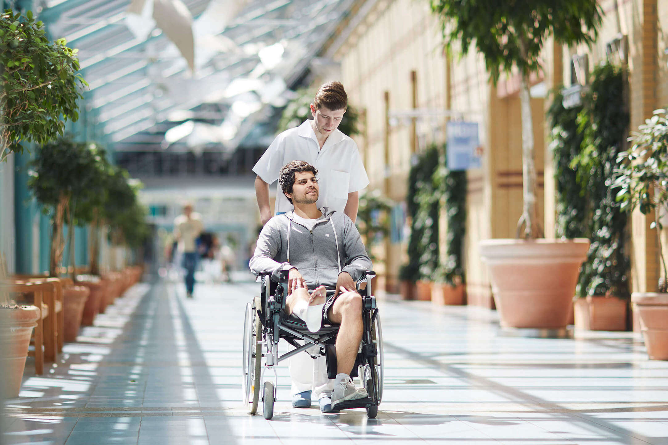 Ein Pfleger schiebt einen Patienten mit Gips am Bein im Rollstuhl